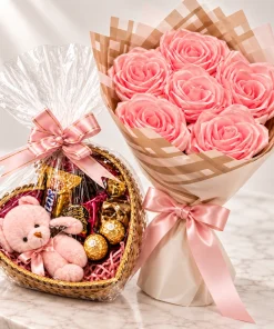 Buquê de rosas de cetim rosa ao lado de cesta em formato de coração com bombons e ursinho