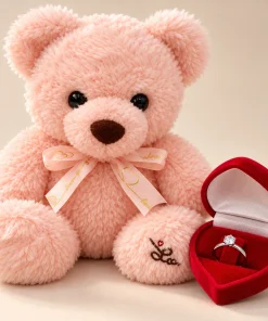 Urso de pelúcia rosa de 20cm com laço decorativo ao lado de caixinha de coração vermelha com anel solitário em aço cirúrgico