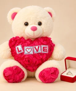 Urso de pelúcia creme de 30cm segurando coração rosa de flores com a palavra LOVE, ao lado de caixinha vermelha com anel solitário
